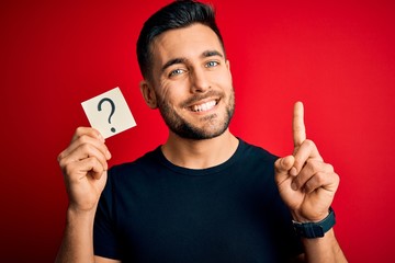 Young handsome man holding paper with question mark symbol over red background surprised with an idea or question pointing finger with happy face, number one