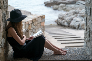 Girl reads book. A girl in a black hat and black silk dress sits in the niche of a fishing hut on the seashore and reads a book. Ocean view at a background. Studying outdoors, reading a novel.