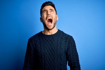 Young handsome man wearing casual sweater standing over isolated blue background angry and mad screaming frustrated and furious, shouting with anger. Rage and aggressive concept.