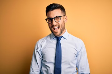 Young handsome businessman wearing tie and glasses standing over yellow background winking looking...