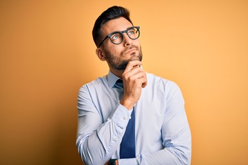 Young handsome businessman wearing tie and glasses standing over yellow background with hand on chin thinking about question, pensive expression. Smiling with thoughtful face. Doubt concept.