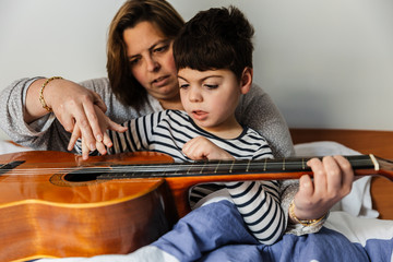 niño juega y aprende con  una mujer a tocar la guitarra en la cama. son madre e hijo, él tiene...
