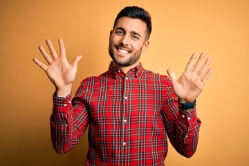 Young handsome man wearing casual shirt standing over isolated yellow background showing and pointing up with fingers number ten while smiling confident and happy.