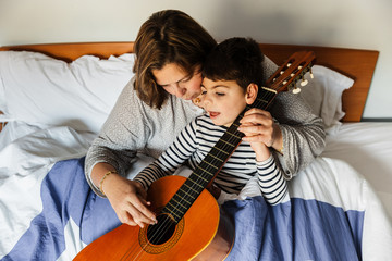 niño juega y aprende con  una mujer a tocar la guitarra en la cama. son madre e hijo, él tiene...