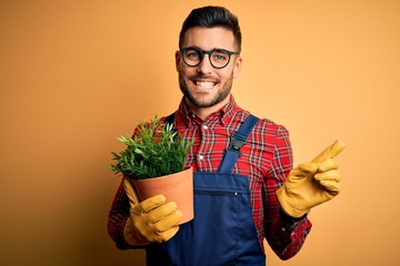 Young gardener man wearing working apron gardening plat for hobby over yellow background very happy...