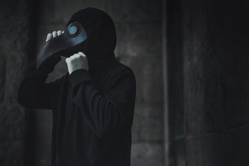 Dark horror figure in black hood in plague doctor mask on the grunge background. Stylized epidemic costume