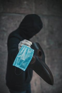 A man in black hood holds a mask of plague doctor and disposable medical mask. Epidemic protection