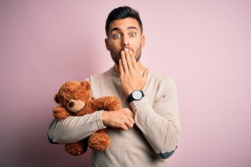 Young handsome man holding teddy bear standing over isolated pink background cover mouth with hand shocked with shame for mistake, expression of fear, scared in silence, secret concept