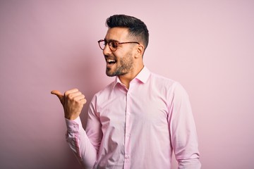 Fototapeta premium Young handsome man wearing elegant shirt and glasses standing over pink background smiling with happy face looking and pointing to the side with thumb up.