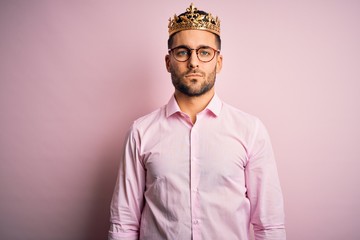Obraz premium Young handsome business man wearing golden crown as king over pink background with serious expression on face. Simple and natural looking at the camera.
