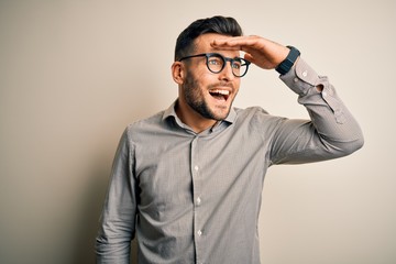 Fototapeta premium Young handsome man wearing elegant shirt and glasses over isolated white background very happy and smiling looking far away with hand over head. Searching concept.