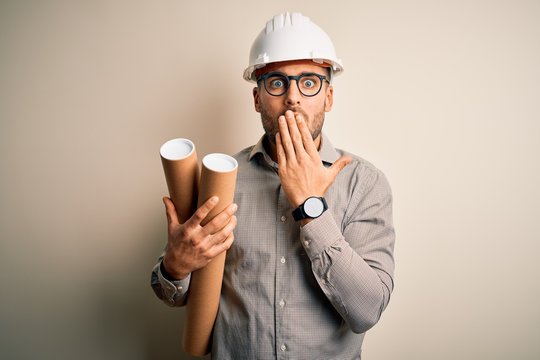 Young architect man wearing contractor helmet holding project paper plan over isolated background cover mouth with hand shocked with shame for mistake, expression of fear, scared in silence, secret