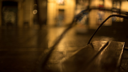 Rainy evening in the city. Shiny raindrops on a the bench. Multi-colored city lights in the background. Creative evening lights and blurred shadows. Twilight mood. Close up.