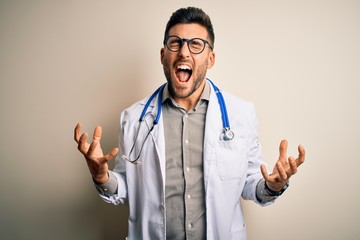 Young doctor man wearing glasses, medical white robe and stethoscope over isolated background crazy...