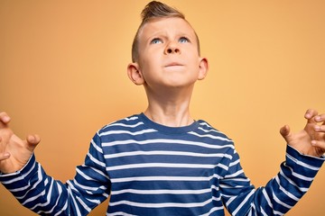 Young little caucasian kid with blue eyes wearing nautical striped shirt over yellow background crazy and mad shouting and yelling with aggressive expression and arms raised. Frustration concept.