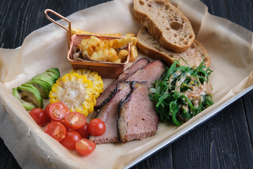 Grilled steaks with vegetables, corn, tomatoes, lettuce, French fries, slices of bread on a rectangular platter for serving on a wooden background