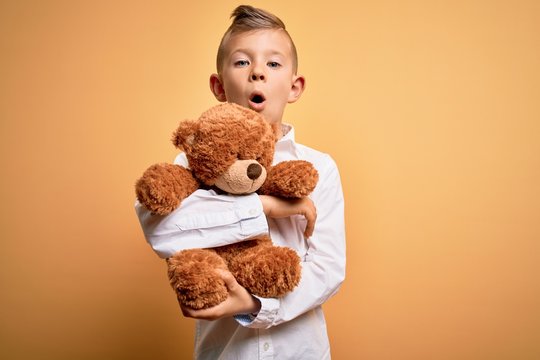 Young little caucasian kid hugging teddy bear stuffed animal over yellow background scared in shock with a surprise face, afraid and excited with fear expression