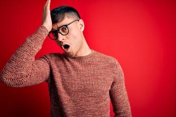 Young handsome hispanic man wearing nerd glasses over red background surprised with hand on head...