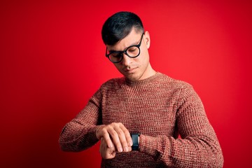 Young handsome hispanic man wearing nerd glasses over red background Checking the time on wrist...