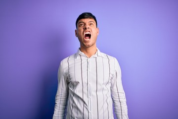 Young handsome hispanic man wearing elegant business shirt standing over purple background angry and mad screaming frustrated and furious, shouting with anger. Rage and aggressive concept.