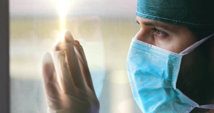 Medico o infermiere uomo con cuffia e mascherina guarda stanco fuori dalla finestra.