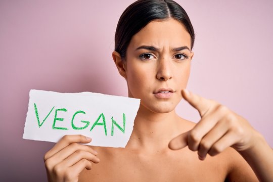 Young beautiful girl holding paper with vegan message over isolated pink background pointing with finger to the camera and to you, hand sign, positive and confident gesture from the front