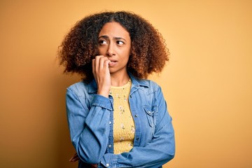 Young african american woman with afro hair wearing casual denim shirt over yellow background looking stressed and nervous with hands on mouth biting nails. Anxiety problem.