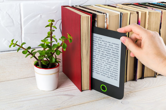 Woman hand takes e-reader from shelf with paper books and small potted plant. Copy space on e-book display. E-reading for pleasure and education.