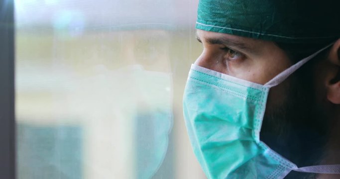Medico o infermiere uomo con cuffia e mascherina guarda stanco fuori dalla finestra.