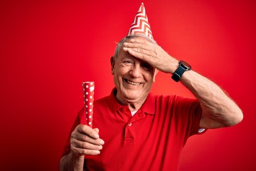 Senior grey haired man wearing birthday hat and holding party trumpet over red background stressed with hand on head, shocked with shame and surprise face, angry and frustrated. Fear and upset