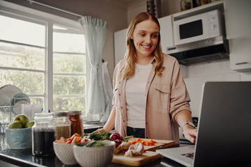 Cheerful young female blogger browsing for online recipe to prepare salad of fresh vegetables and...
