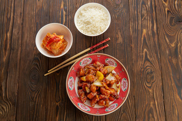Asian cuisine, sweet and sour fried pork chunks with bell peppers, onion and pineapple on traditional plate with chopsticks, on old wooden table with rice in white bowl