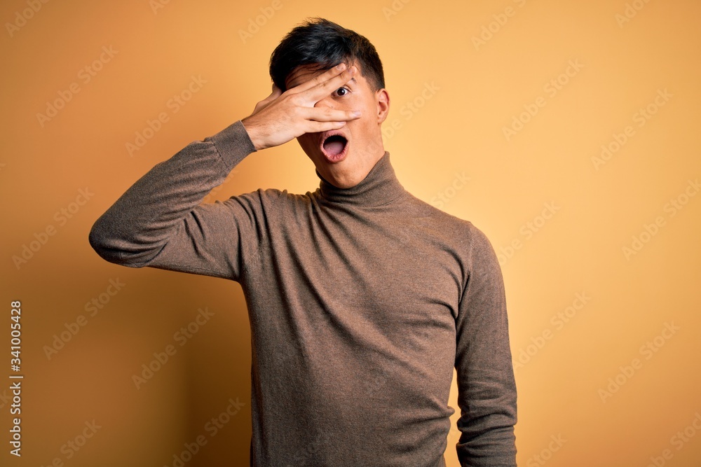 Sticker Young handsome man wearing casual turtleneck sweater over isolated yellow background peeking in shock covering face and eyes with hand, looking through fingers with embarrassed expression.