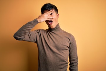 Young handsome man wearing casual turtleneck sweater over isolated yellow background peeking in shock covering face and eyes with hand, looking through fingers with embarrassed expression.