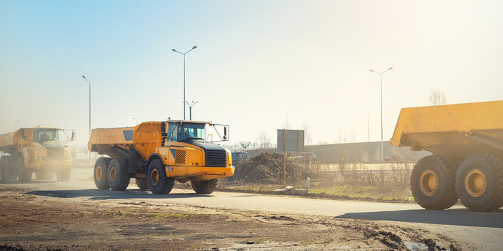 Many big articulated heavy industrial yellow dumper trucks driving on new highway road construction site on sunny day with blue sky background. Construction equipment machinery working on open pit