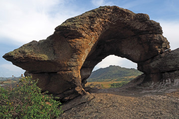 The fantastic stone arch on the big brown rock with spots of lichens, the green bush near it, the blue sky with white clouds