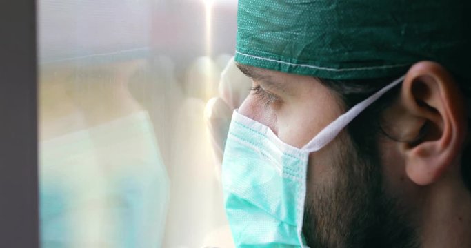 Medico o infermiere uomo con cuffia e mascherina guarda stanco fuori dalla finestra.