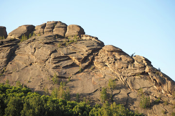 The fantastic high rock with cracks and the birch forest at the its foot, the clean blue sky in the summer evening