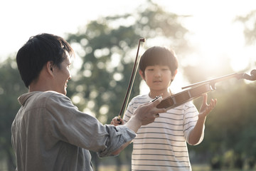 Asian father give and teach young boy kid to play violin in park together. Man educate music skill to his child to be specialist and professional in future. Nature outdoor violin class in evening. © Kawee