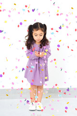 Obraz na płótnie Canvas Beautiful small girl blowing colorful confetti. Portrait of a little child wearing dress standing under confetti rain and celebrating over white background.