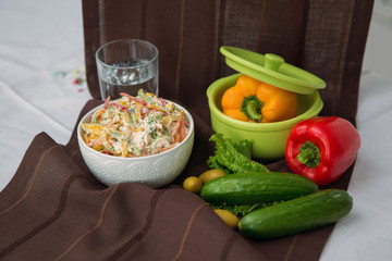 Decorated with lettuce, side view, selective focus . Orange pepper in a green bowl . Water in a glass. Brown tablecloth. Salad with ham, bell pepper, mushrooms, pickles, white sauce, on a white plate.