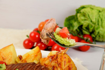 grilled meat on a fork, pieces of grilled potato next to lettuce with tomatoes and olives on a white plate