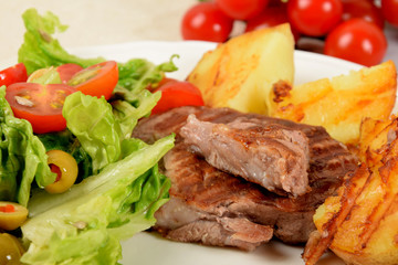 pieces of grilled steak next to potatoes grilled next to lettuce with tomatoes and olives on a white plate