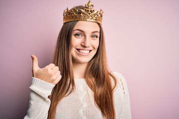 Young beautiful redhead woman wearing queen crown over isolated pink background doing happy thumbs...