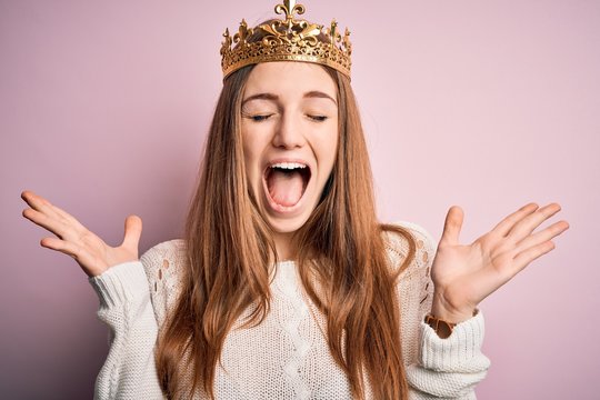 Young beautiful redhead woman wearing queen crown over isolated pink background celebrating mad and crazy for success with arms raised and closed eyes screaming excited. Winner concept