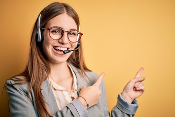 Young redhead call center agent woman overworked wearing glasses using headset smiling and looking at the camera pointing with two hands and fingers to the side.