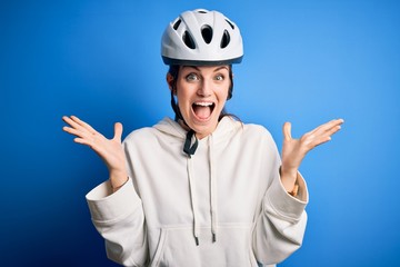 Young beautiful redhead cyclist woman wearing bike helmet over isolated blue background celebrating crazy and amazed for success with arms raised and open eyes screaming excited. Winner concept