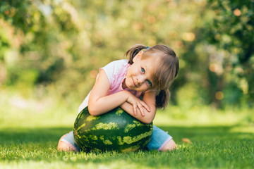 Fototapeta premium Little toddler girl squatting by a large watermelon on the lawn in the summer garden