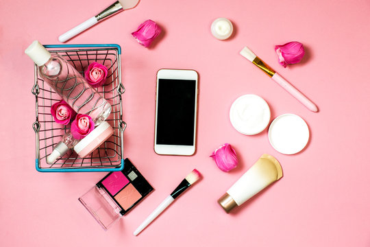 Concept Of Online Shopping Cosmetics. Top View On Cosmetics Bottles, Cream, Soap, Makeup Brushes, Mobile Phone, Credit Card And Shopping Basket From The Supermarket On A Pink Background, Flat Lay