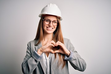 Young beautiful redhead architect woman wearing security helmet over white background smiling in love showing heart symbol and shape with hands. Romantic concept.